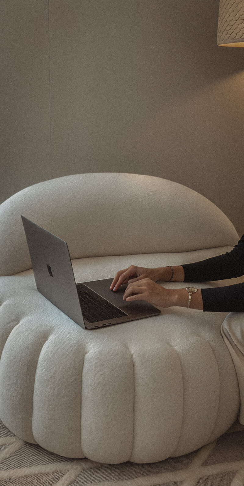 Black-haired woman working on a laptop while seated on a modern white boucle chair in a neutral-toned, softly lit space — calm and minimal workspace aesthetic