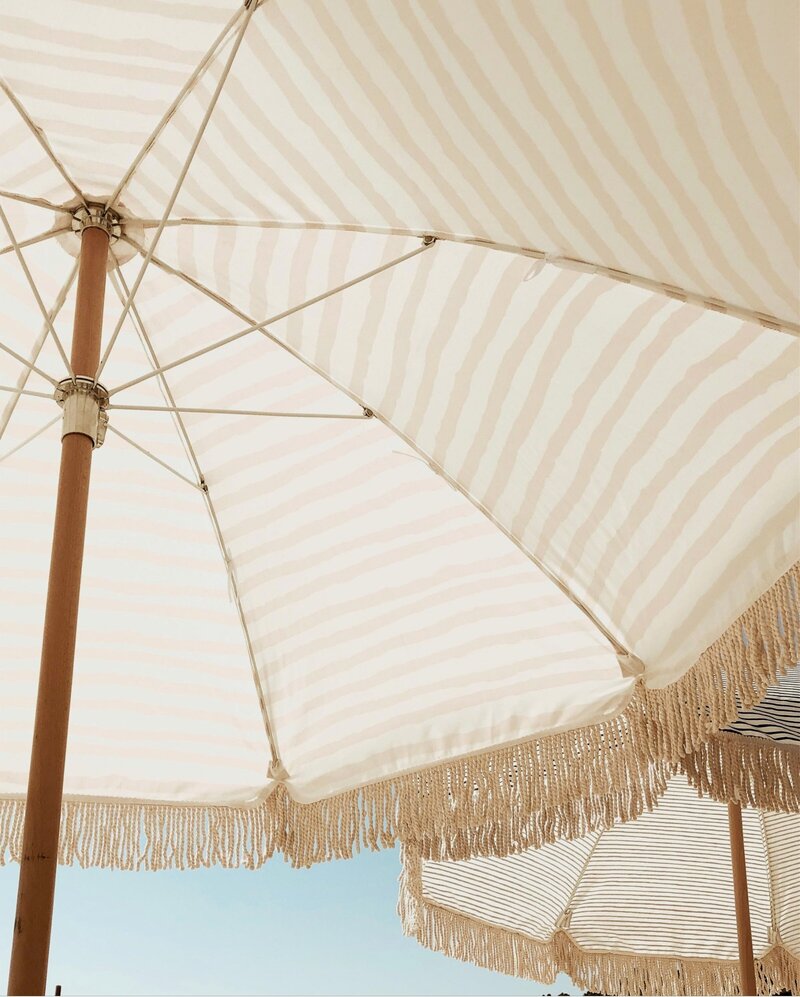 Coastal sun umbrellas in tan stripes providing shade on a sunny day with blue sky backdrop. Tailored Oasis Travel Co.