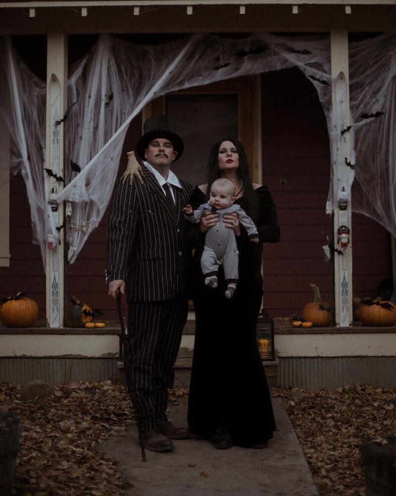 A family posing in front of their decorated home for Halloween. They are in Adams family costumes. The mother is holding her child as the father stands next to them with a cain and top hat