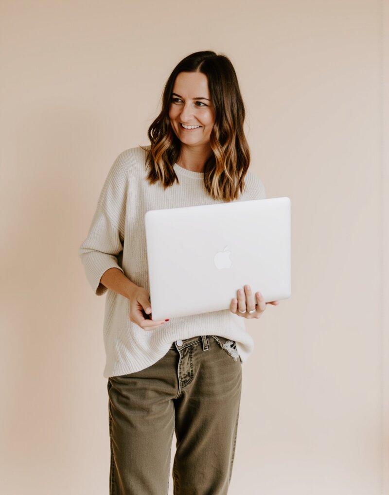 A woman wearing a white sweater and olive pants is holding a laptop, looking focused and engaged.