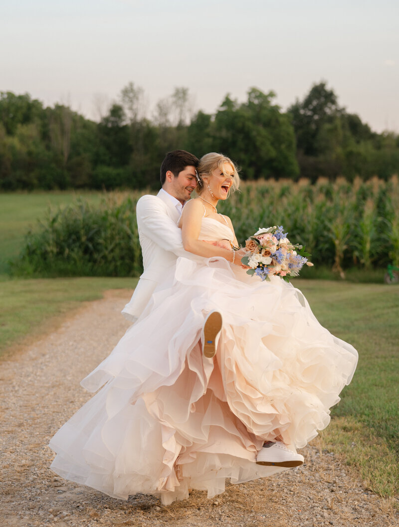 A groom in white picks up his bride in pale pinks and spins her around at golden hour 