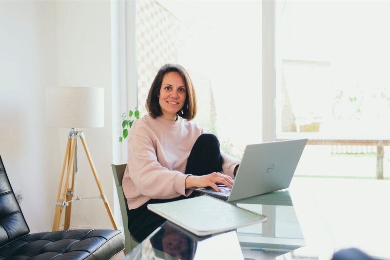 Kaya sitting at a glass desk with a laptop, smiling toward the camera in a bright room