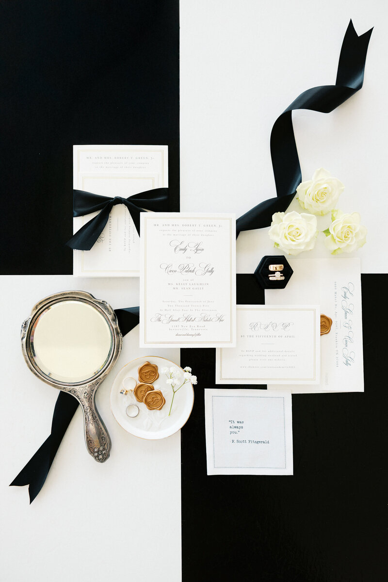 Flatlay of black and white wedding invitation suite with wax seals, vintage mirror, wedding rings, and white roses at Nichols Heir in Tennessee
