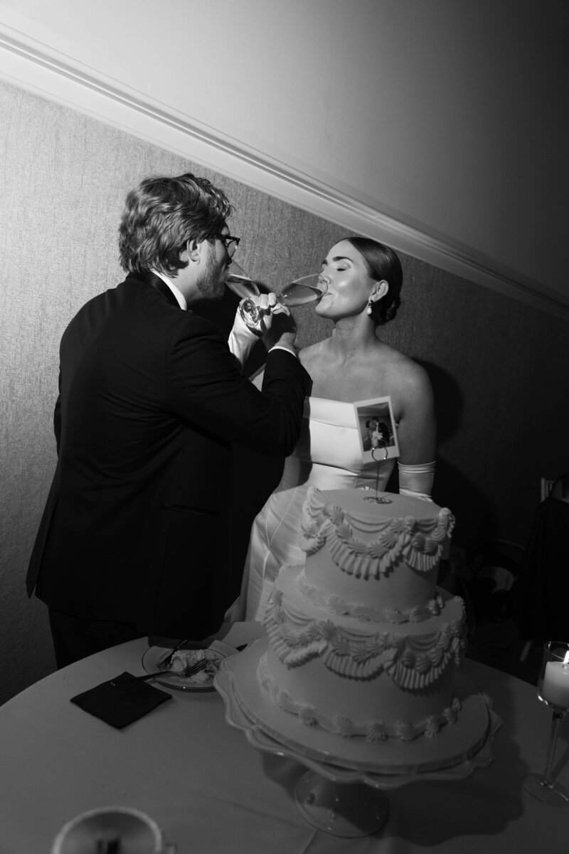 Candid moment of newlyweds sharing cake together at reception, classic black and white editorial wedding photo.