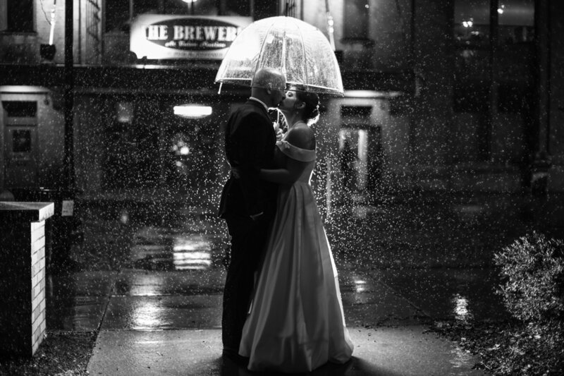 Bride and groom kiss under an umbrella during a rainstorm at night in front of the Brewerie at Union Station in Erie PA