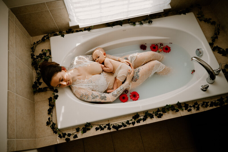 Motherhood photo looking down at mom in bathtub laying back in white while nursing baby girl with pink flowers around them in Annapolis Maryland.