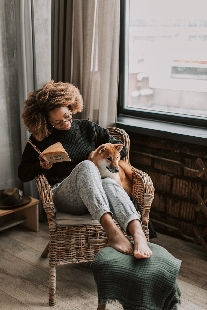 This is a picture of a black woman sitting in a chair with her dog cuddling and reading.