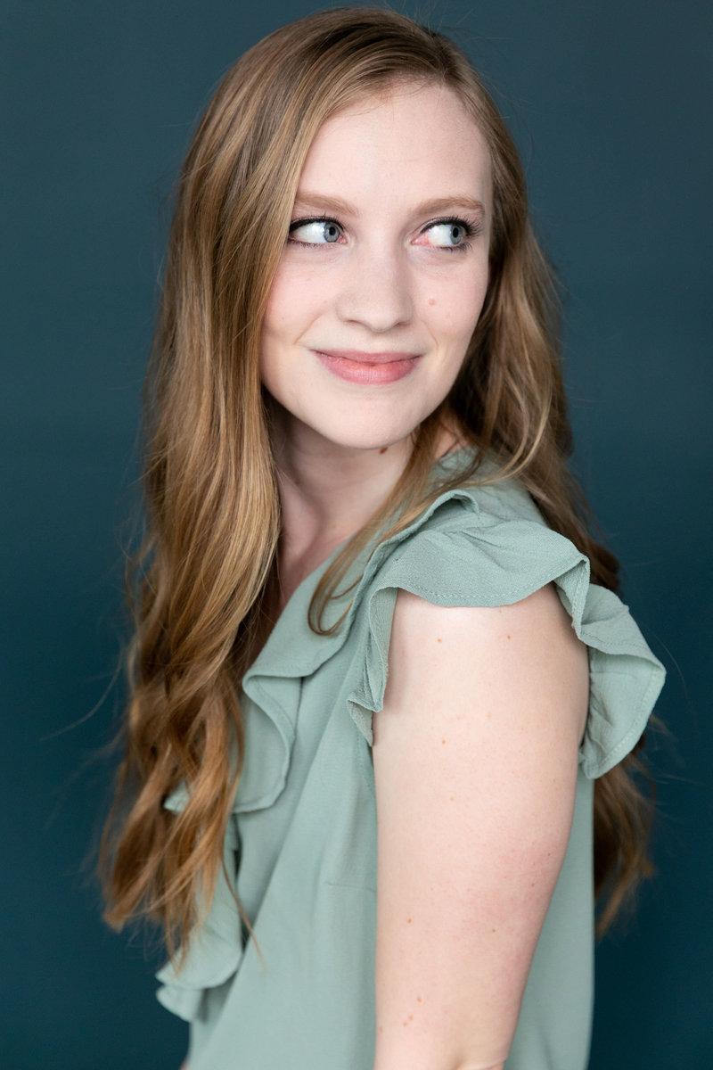 Blonde woman in a ruffled, teal shirt peers to the side with a smile on her face