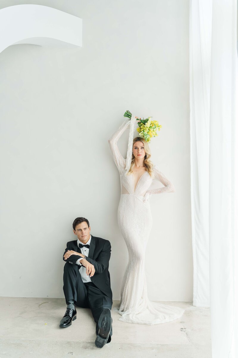 A couple on their wedding gowns, the bride is standing while holding the bouqete of yellow flowers over her head and the groom is sitting down on the floor