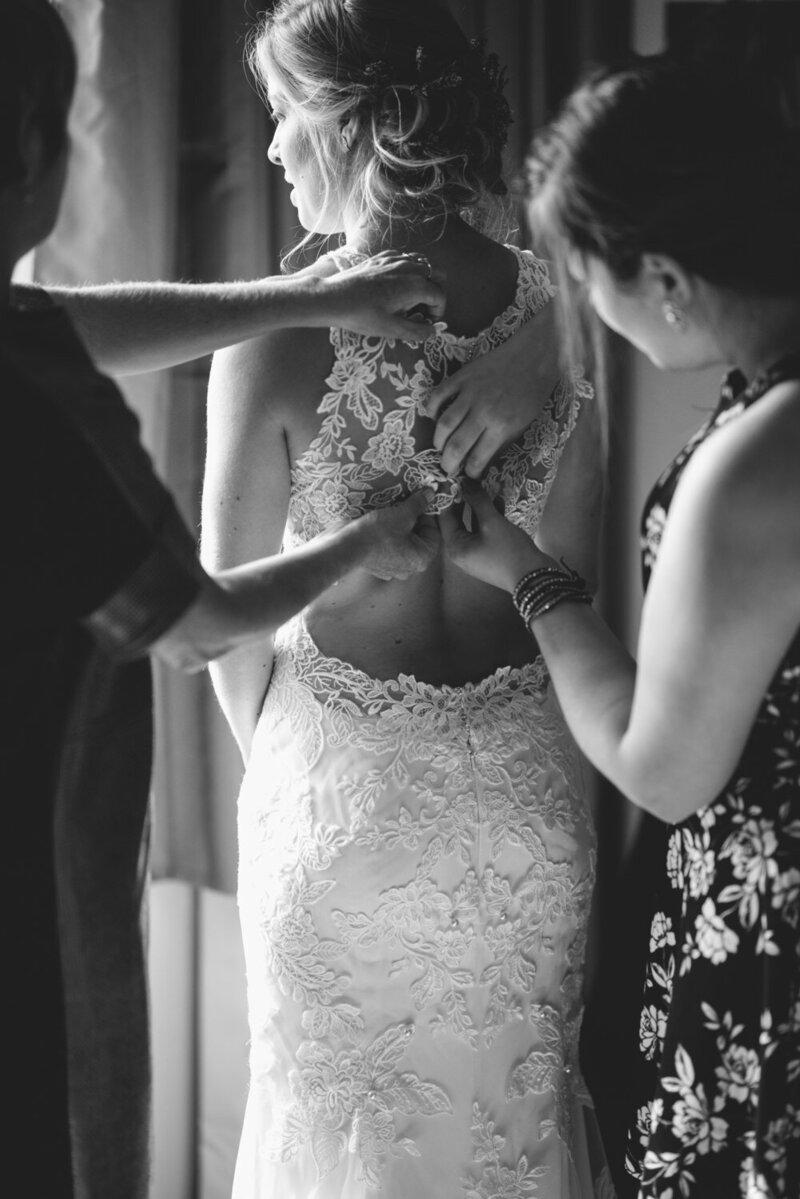 La mariée finalise sa préparation entourée de ses demoiselles d’honneur qui l’aident à attacher sa robe, moment complice et touchant capturé dans l’intimité d’un mariage élégant et chaleureux