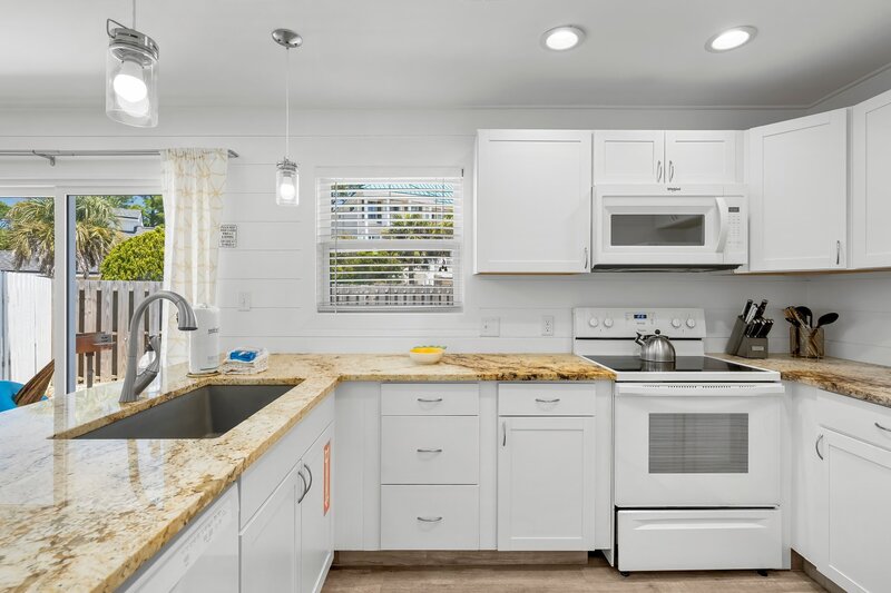 Modern white kitchen with granite countertops, full-size appliances, and a window view of palm trees and tropical surroundings.
