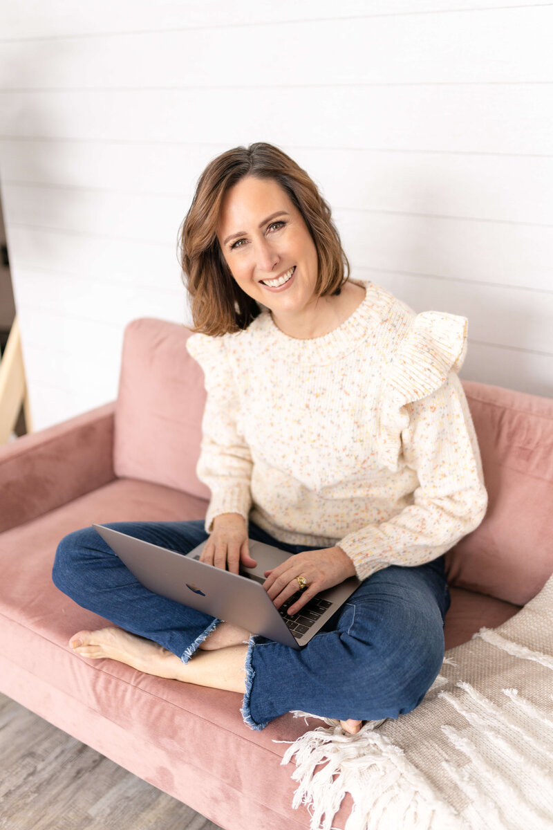 Female writer sitting on pink couch typing on laptop and smiling.