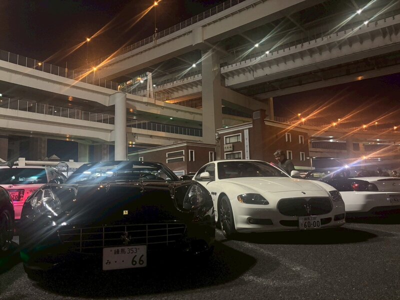 A Ferrari and a Maserati parked side by side under the illuminated expressways at Daikoku Parking Area during the night.