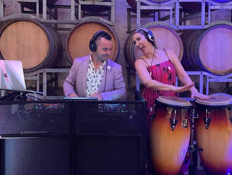 Wedding percussionist performing on congas next to a DJ at a winery venue with stacked barrels.