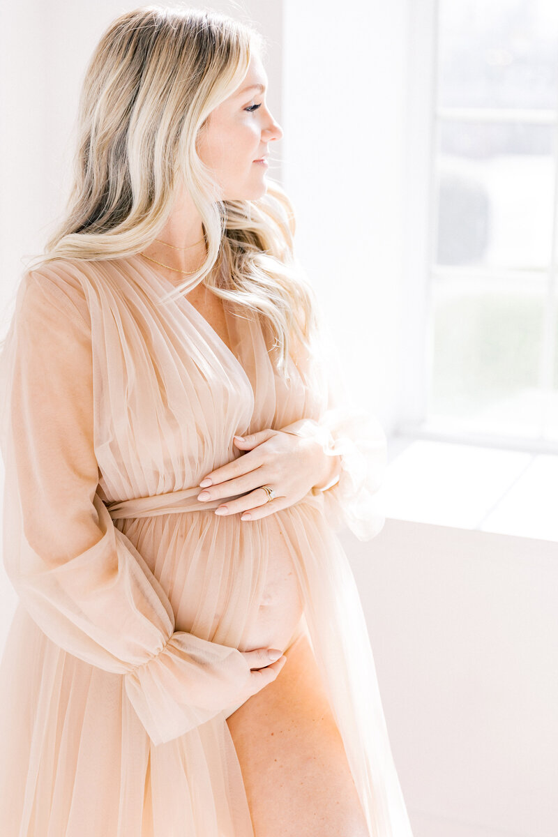 A mom to be stands smiling out a window in a pink maternity gown with hands on bump