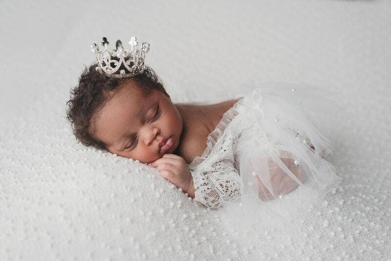 Newborn baby girl wearing crown and white lace onesie with lace tutu  in all white against white backdrop with texture
