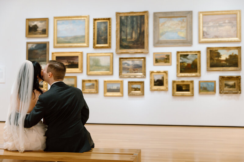 Bride and groom sitting on a bench in the Akron Art Museum with a gallery of paintings behind them. 