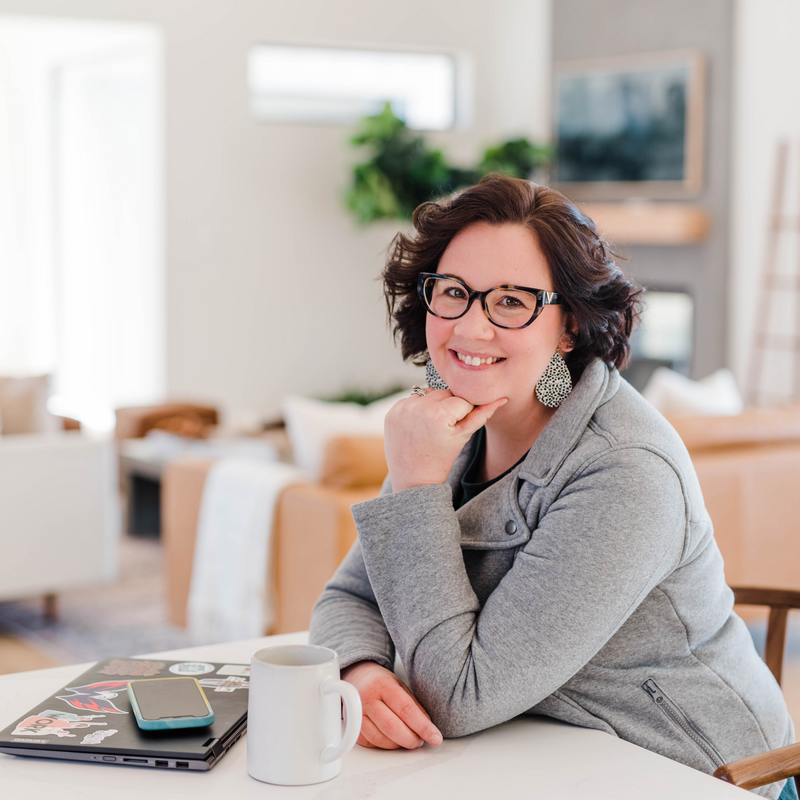 A woman with short, dark hair and glasses smiles while sitting at a table in a cozy, modern living room. She rests her chin on her hand, with a laptop, smartphone, and coffee mug on the table in front of her. The room features soft lighting and a neutral color palette, with greenery and a wall-mounted television in the background.