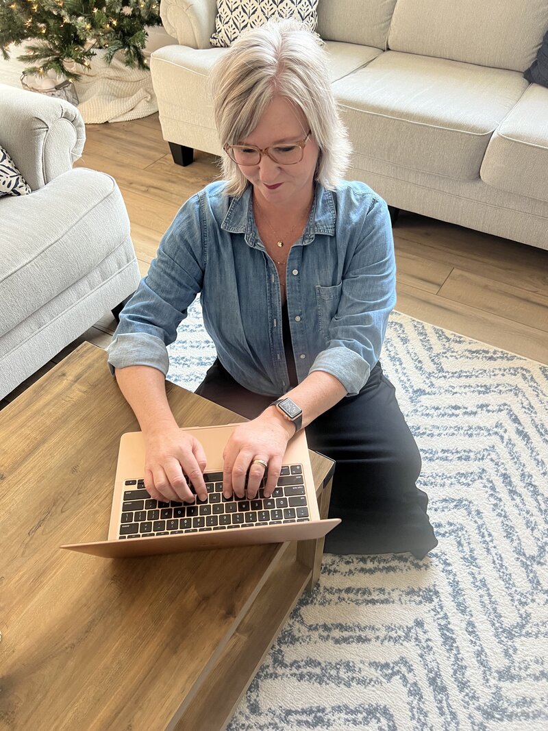 Jenny Laine Designs sitting on the floor in a living room at a laptop placed on a coffee table.