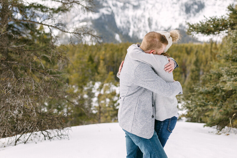 This warm image features a fiancée hugging her fiancé from behind among the snowy woods of Lake Louise. Their smiles reflect a joyful connection, capturing a tender moment of love and laughter in the serene, wintry forest setting.