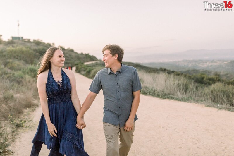 Engaged couple smile as they walk hand in hand along a dirt hiking trail