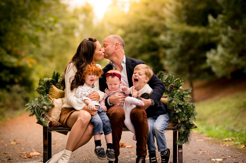 Parents kissing during a holiday family photoshoot session with their three children. Photograph shows a beautiful glowy, misty backdrop along a private path in Redmond WA.