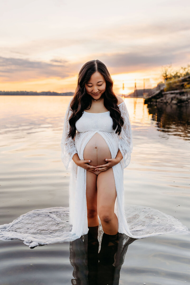 A pregnant mom stands in a river at golden hour, looking at her pregnant belly and smiling, during her maternity session in Vancouver Washington