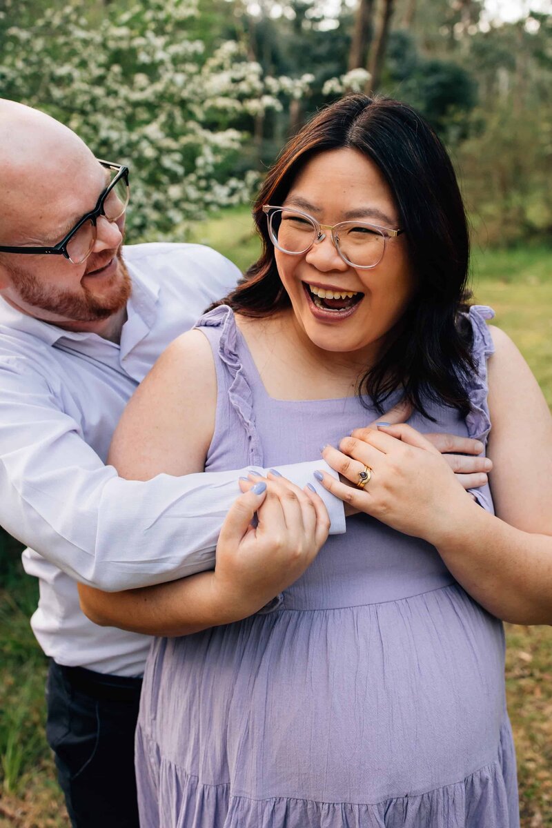 pregnant women wearing red maternity dress being embraces by husband's arms