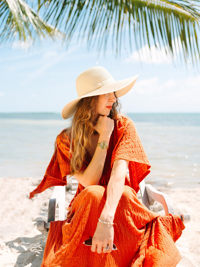 Kilee in an orange dress and straw hat sitting in chair on the beach with ocean behind her