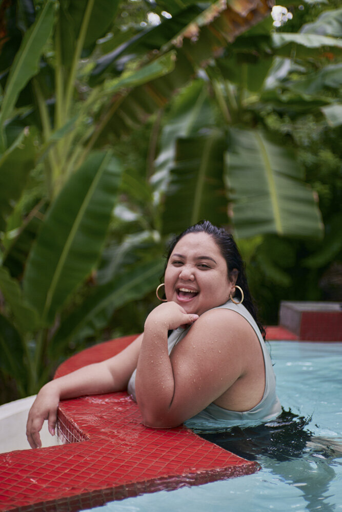 A plus-size woman of color smiles brightly toward the camera, her chin resting in one hand, leaning over the edge of a pool. She is wearing gold hoop earrings, with long hair down her back. Large tropical plants are visible in the background of the image.