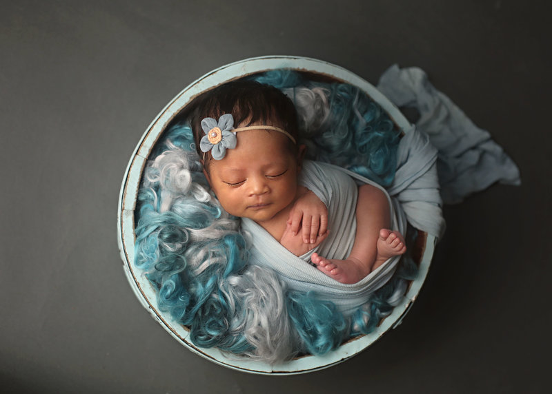 newborn in light blue wraps posed in a basket filled with different shades of blue blankets