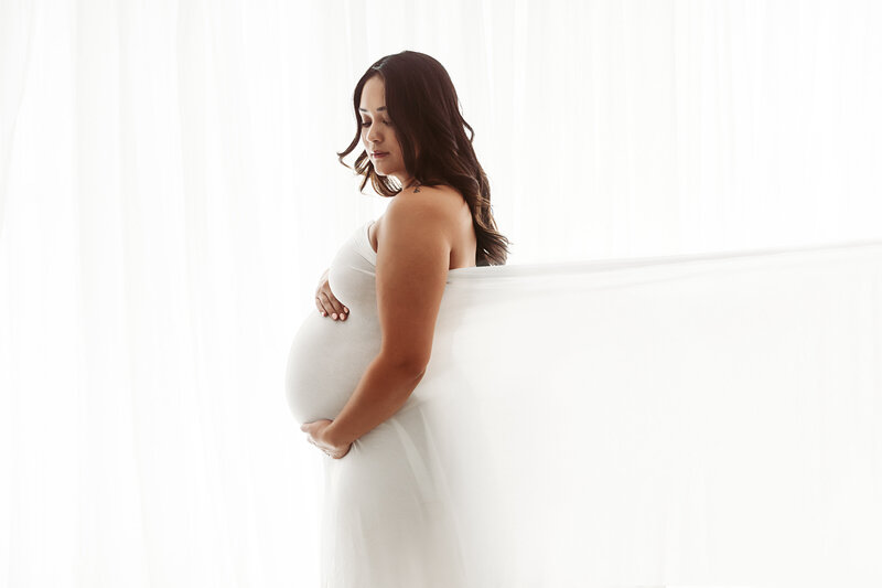 A pregnant woman wearing a white dress stands against a bright, softly lit background. She gently cradles her belly, looking down with a serene expression. Her long, dark hair cascades over her shoulders.