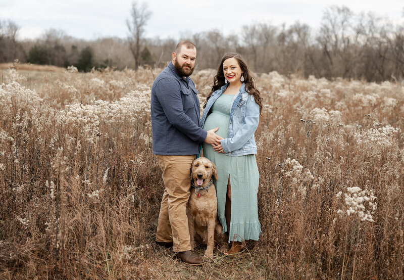 Expecting couple with their dog in the middle standing in a field in Farmington, Missouri. 