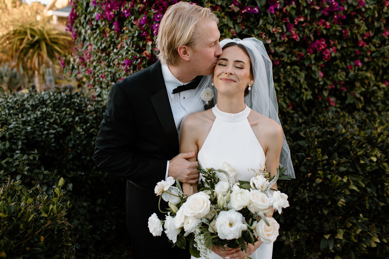 9-radiant-love-event-2021-groom-kissing-brides-head-holding-white-rose-bouquet-purple-flower-bush-backdrop-romantic-elegant-timeless