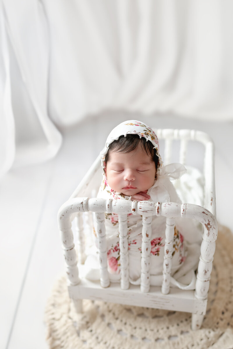 Light, airy vintage crib portrait featuring a newborn girl in a floral bonnet and wrap, bathed in soft white tones.