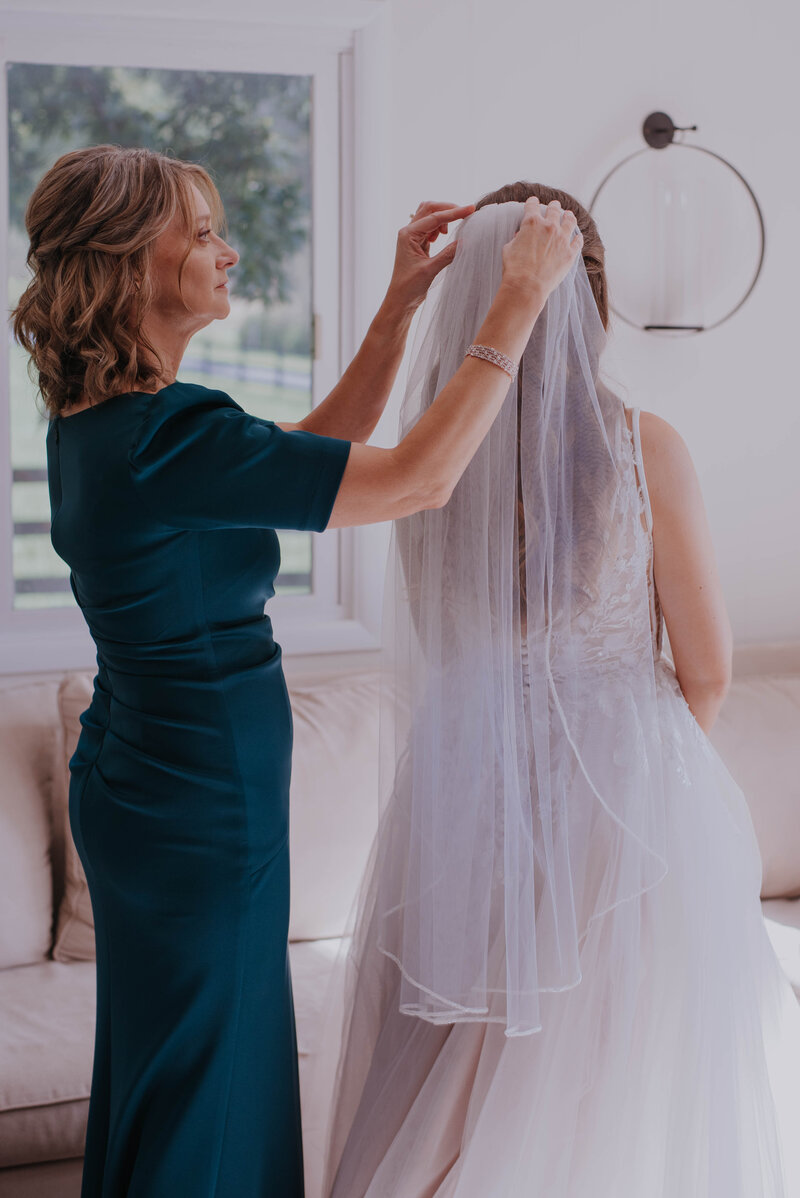 A woman in a dark green dress adjusts the veil of a bride in a white gown. They stand in an elegant reception venue gallery, softly lit, with a beige couch and a window revealing lush greenery outside.
