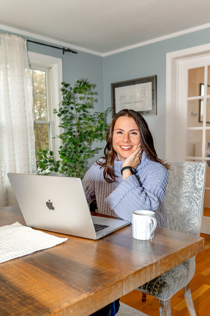 Brunette woman sitting at a wooden dining table, in a blue sweater, smiling  at the camera  while working on laptop with a cup of coffee next to her.