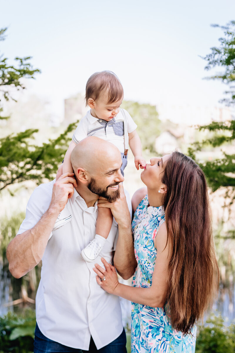 A close-up family portrait featuring a father holding his baby boy on his shoulders while the mother, in a floral dress, playfully interacts with the baby.