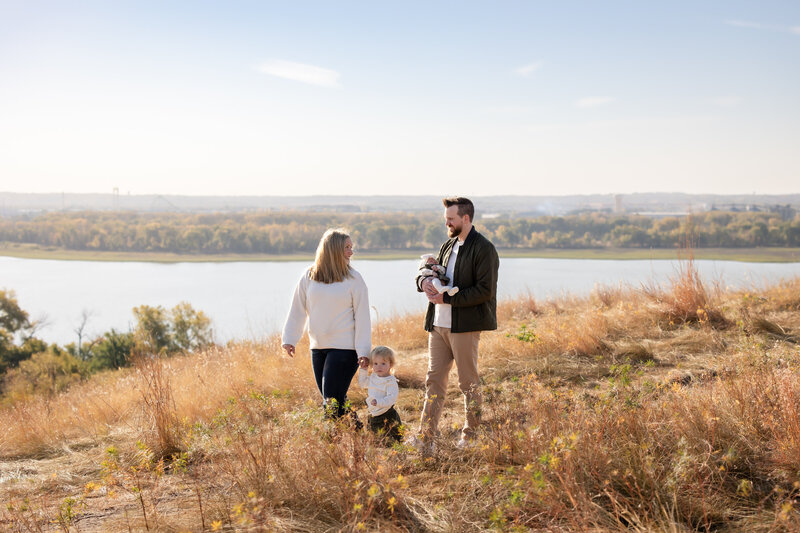 a family walks atop a hillside in Eden Prairie, Minnesota during a fall family photoshoot. A mother holds hands with her daughter as they walk through a field with a lake in the background, mom and dad smile at each other as dad holds a baby.