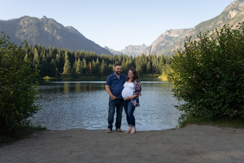 A pregnant couple stand at the edge of gold creek pond in Snoqualmie Pass and laugh together.