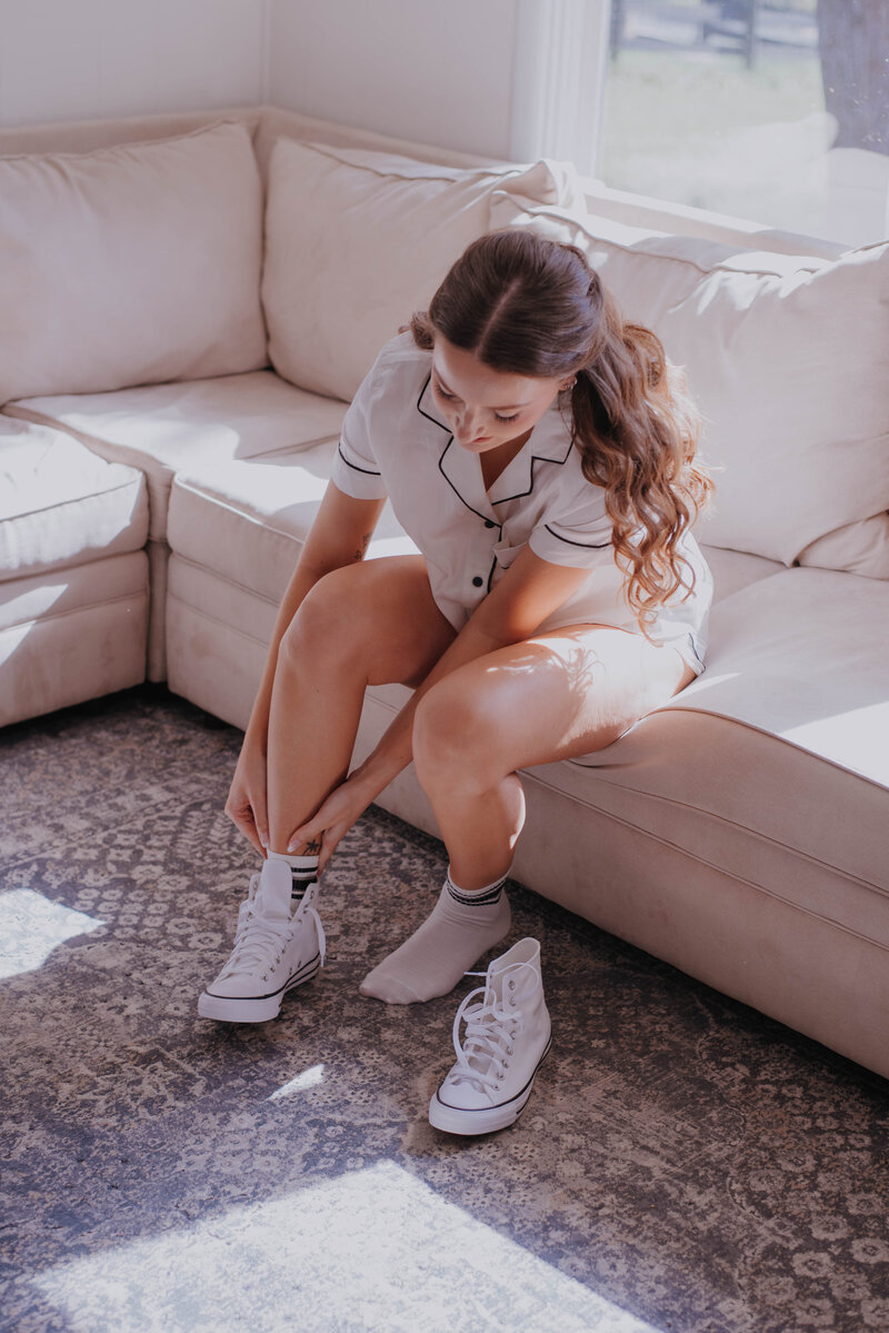 A person lounging on a beige sofa slips on white sneakers over gray socks, dressed in white pajamas with black trim. Sunlight streaming through the window illuminates a patterned rug beneath them, much like the serene ambiance found in outdoor wedding venues throughout Virginia.