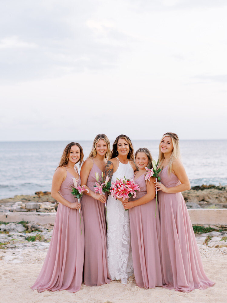 Bride and bridesmaids stand in white and pink dresses holding pink lily bouquets on beach in Mexico for wedding