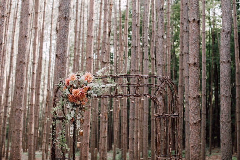 A rustic ceremony arch in the forest featuring peach dahlias and bells