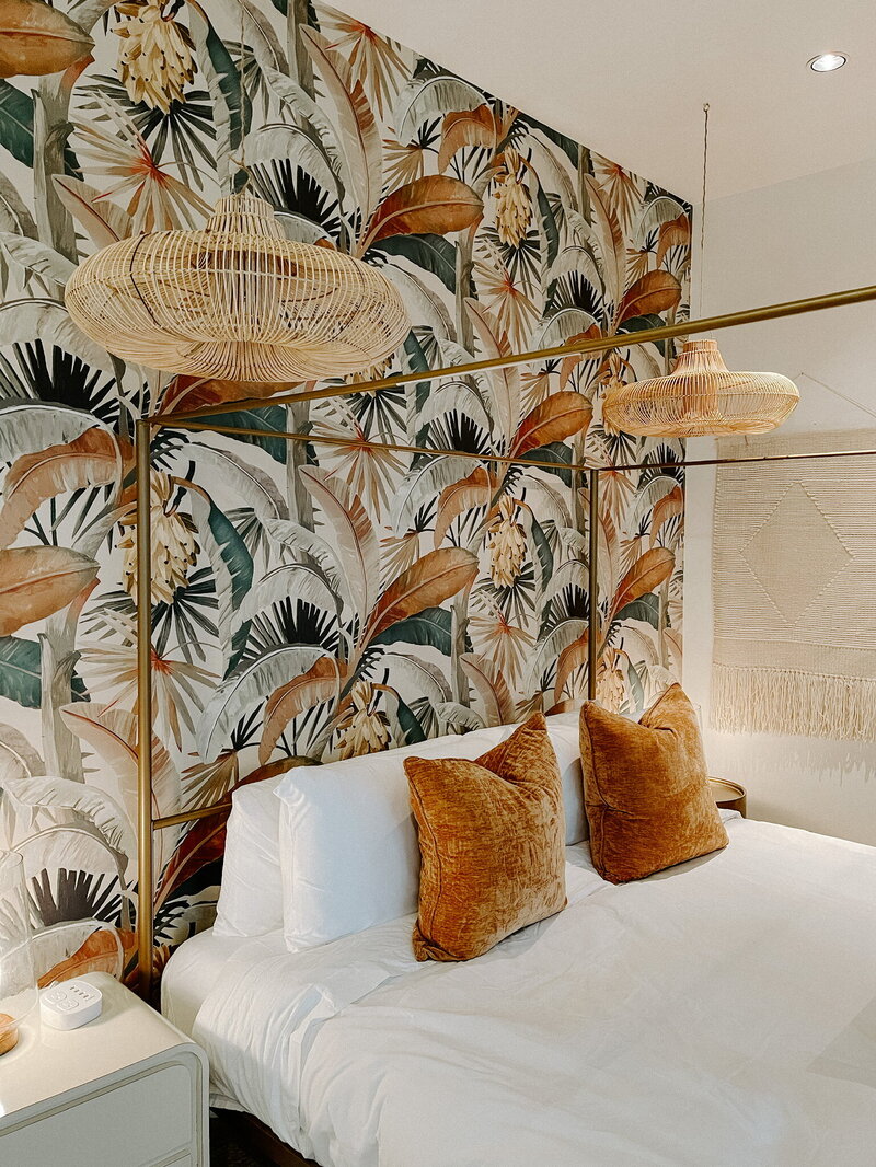 white bed with orange accent pillows against wallpaper of palm leaves