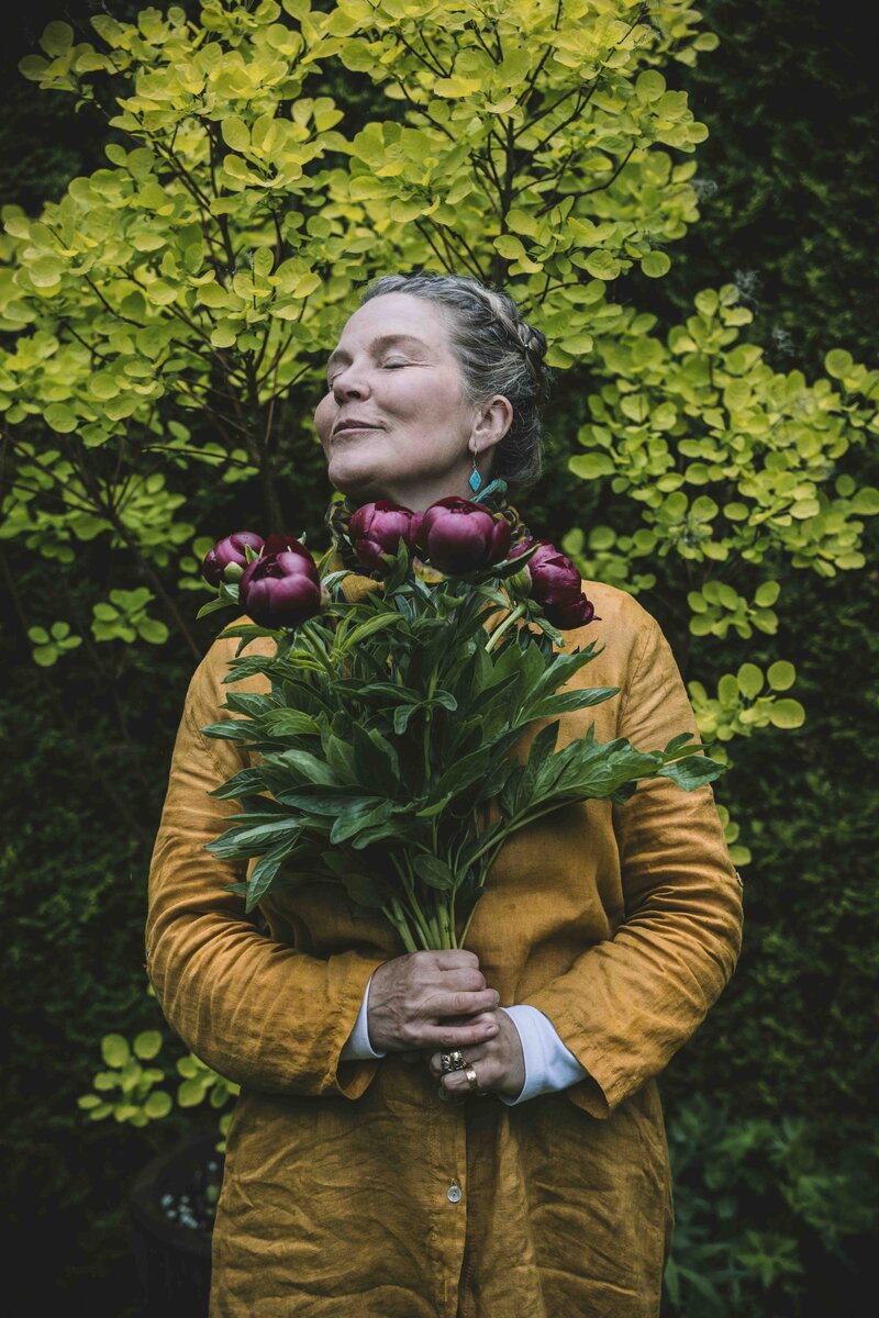 A woman stands gracefully, holding a bouquet of flowers in front of a lush green bush, radiating natural beauty.