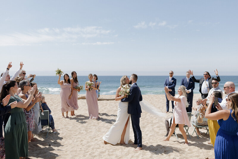 Newlywed couple kissing amidst celebrating guests, candid documentary wedding photography at Montara State Beach.