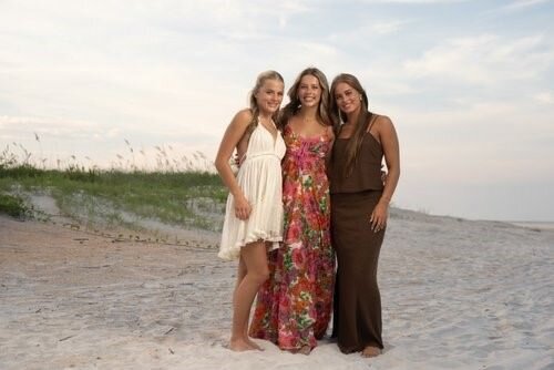 Three-women-in-summer-dresses-stand-on-a-sandy-beach-with-smiles-enjoying-a-serene-day