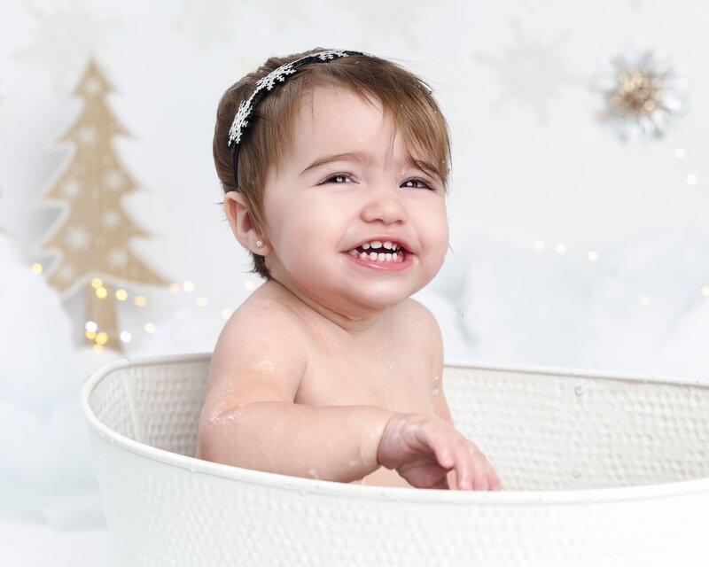 A baby girl with a snowflake headband sitting in a white tub, surrounded by a winter-themed background with snowflakes and a Christmas tree