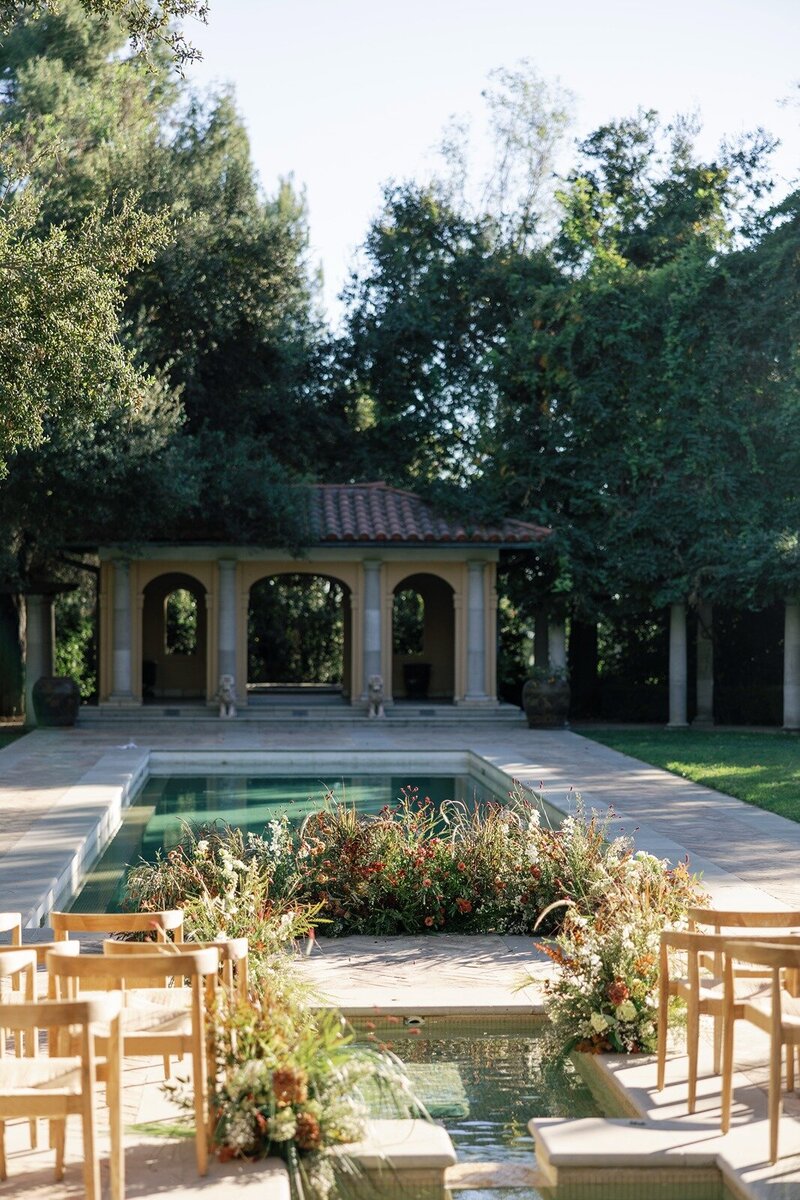 Elegant outdoor wedding ceremony setup featuring wooden chairs, lush floral arrangements, and a reflecting pool at a Mediterranean-inspired estate.