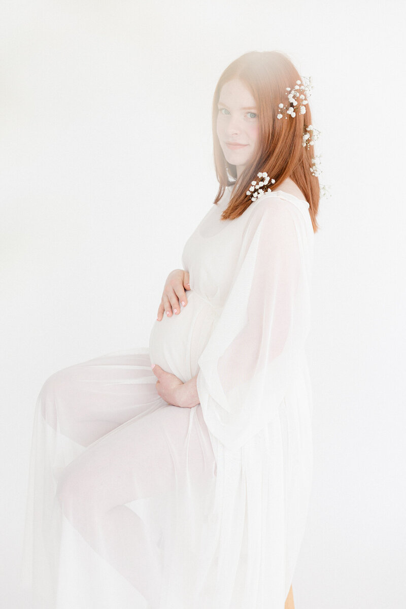 Portrait of an expecting mom wearing a white dress on a white backdrop with flowers in her hair
